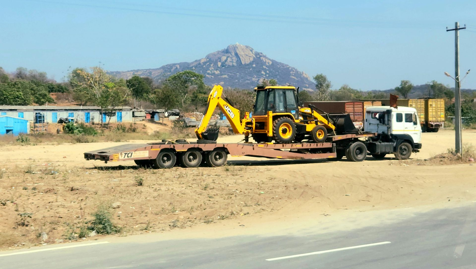 A flatbed truck transports a yellow tractor loader on a sunny day in a landscape with a prominent mountain in the distance.