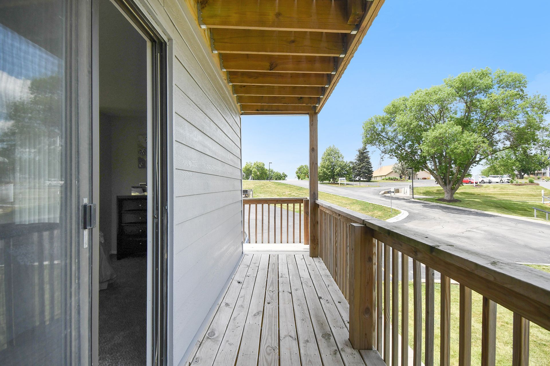 Balcony with wood railing and deck, overlooking a road and trees under a blue sky.