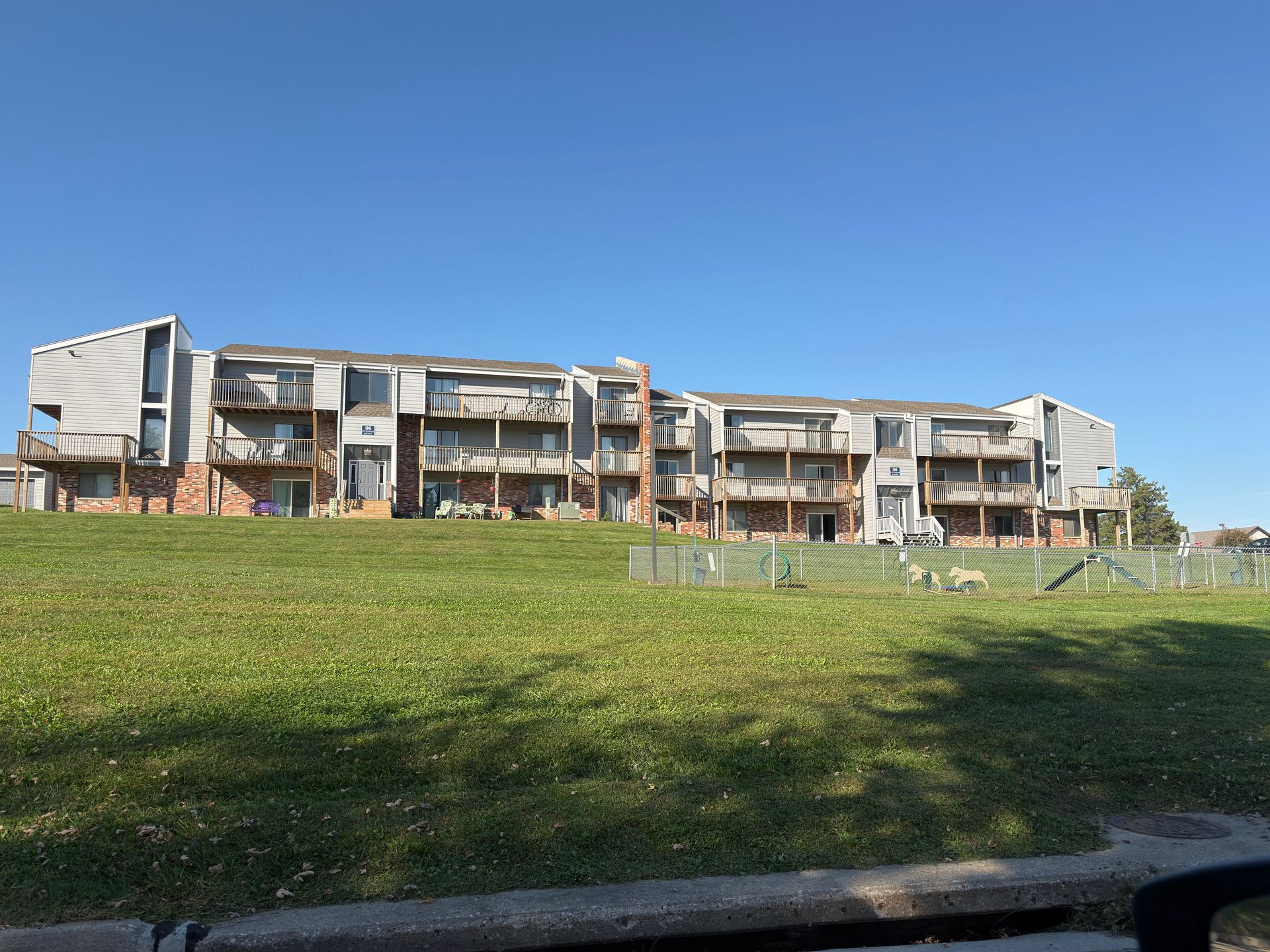 Apartment building on a grassy hill under a clear blue sky. Brown and gray exterior, balconies.
