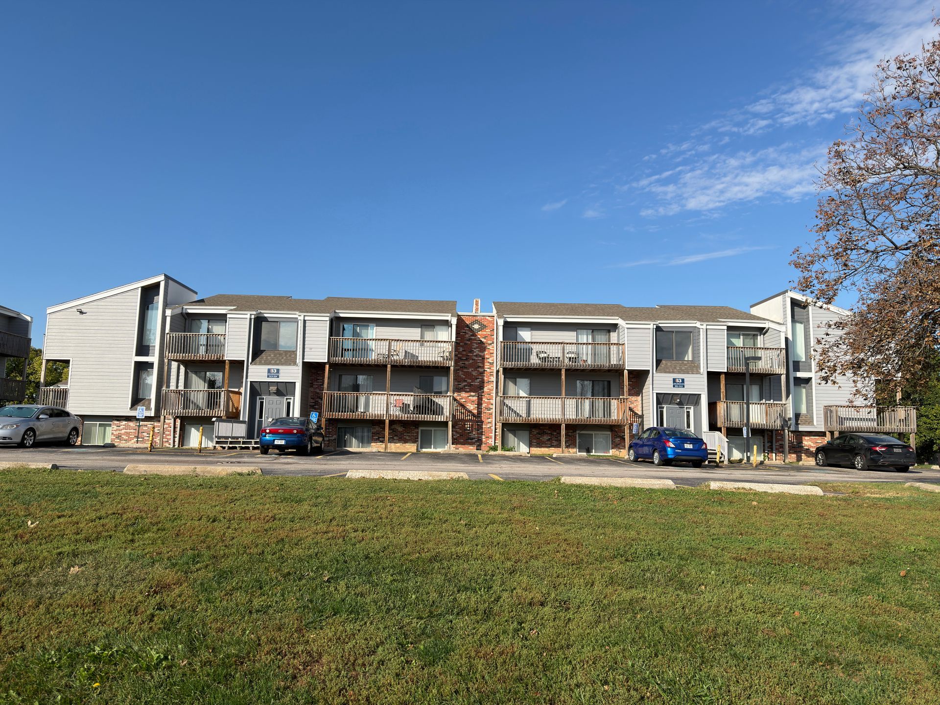 Apartment building exterior, two stories, with balconies and parking spaces in front; blue sky, sunny day.