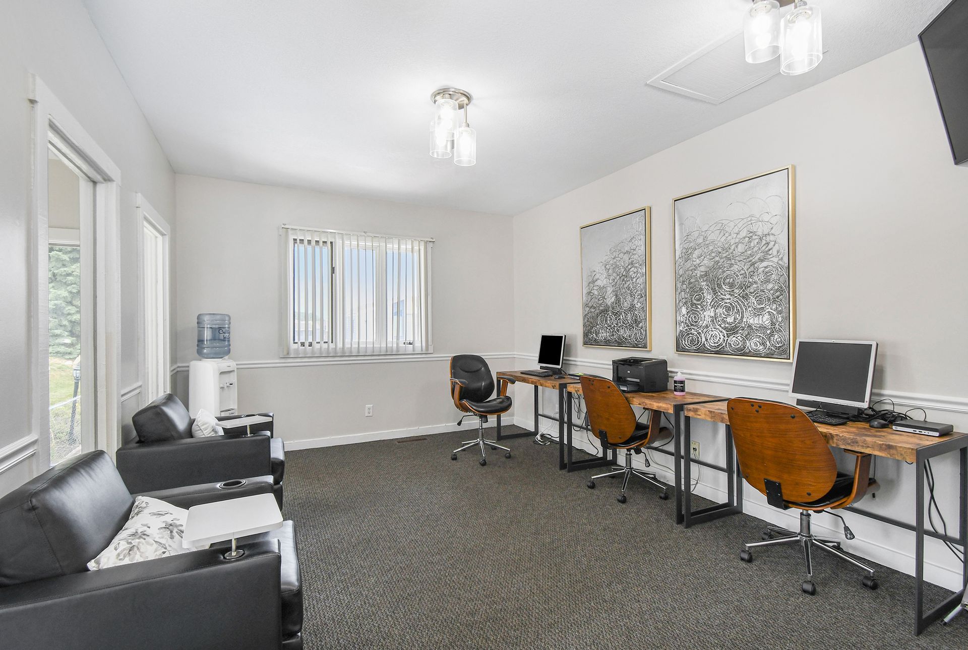 Office space with desks, computers, chairs, and seating area. White walls, grey carpet.