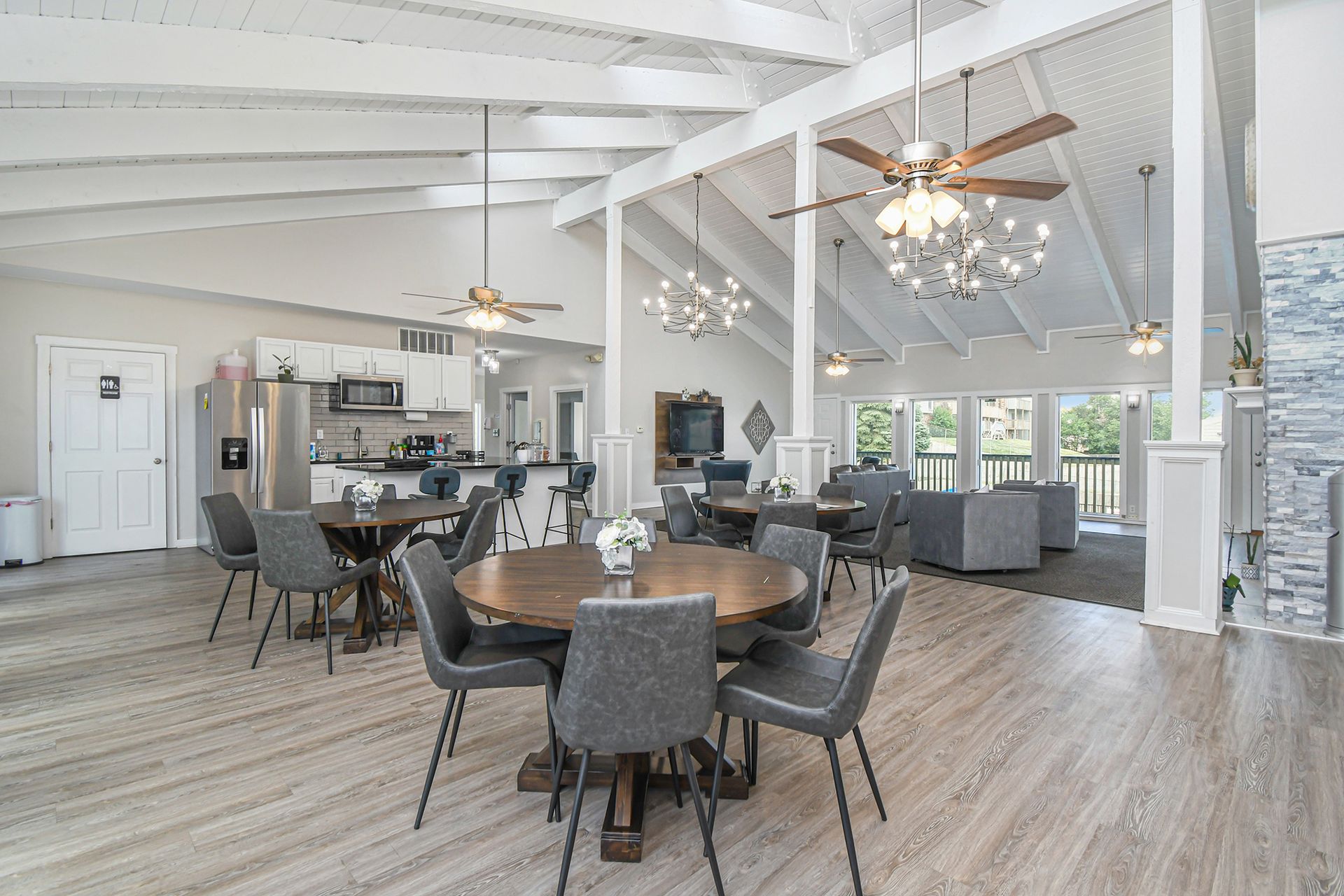Spacious community lounge with dining tables, kitchen, and seating area under a high, white ceiling.