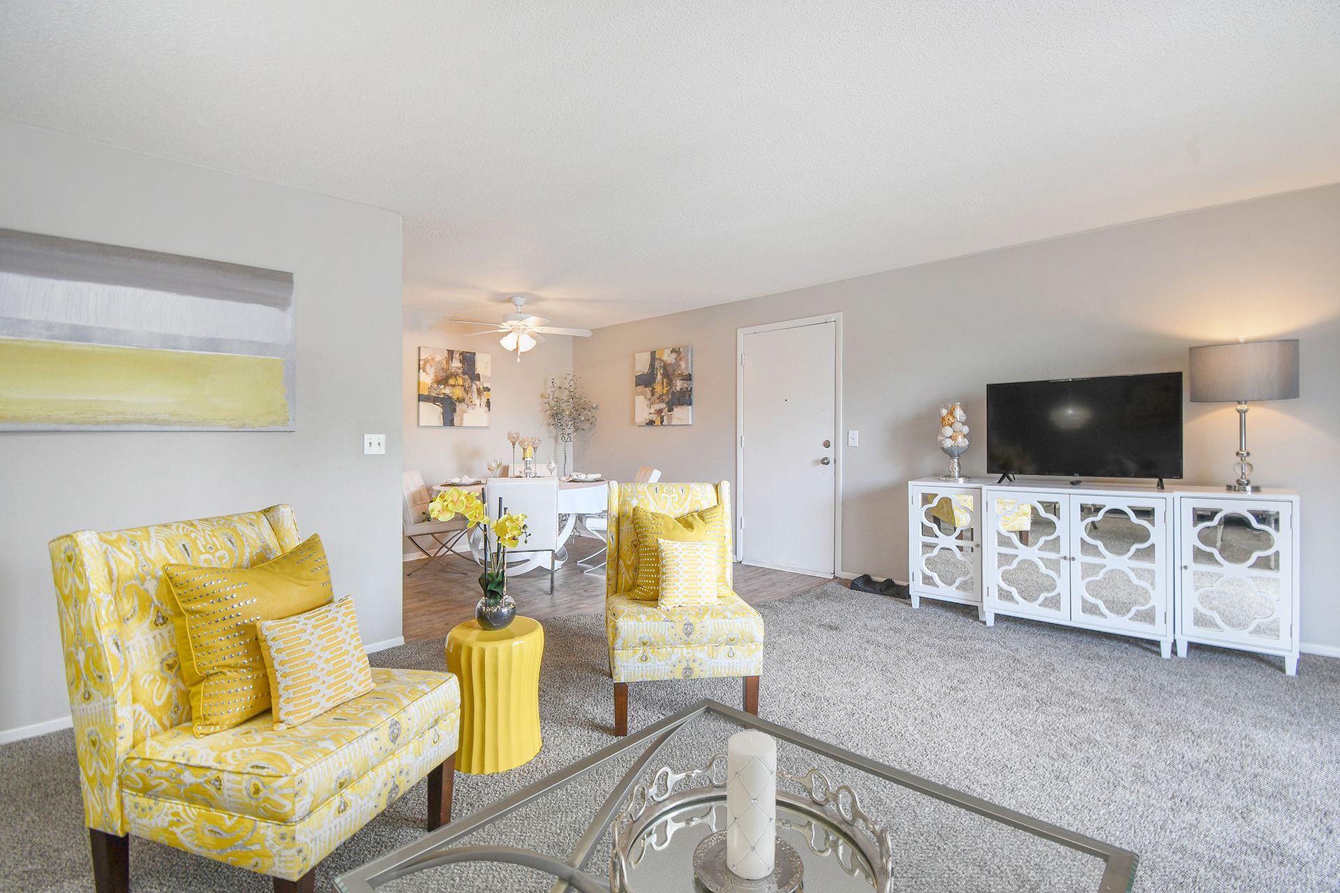 Living room with yellow accents: chairs, pillows, and side table. Gray walls, carpet, and a TV stand.
