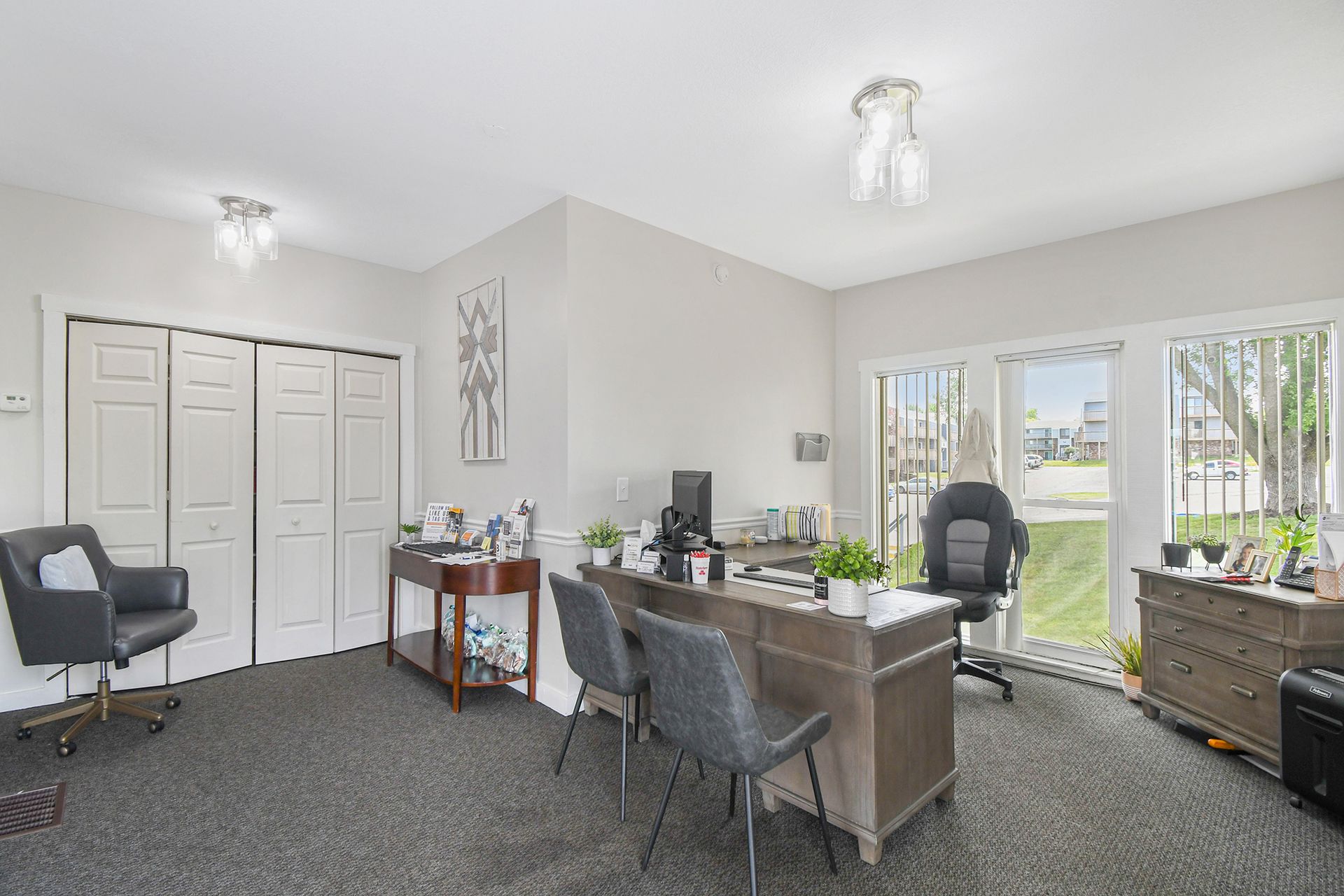 Office with desk, chairs, closed closet doors, window with view of yard. Grey carpet.