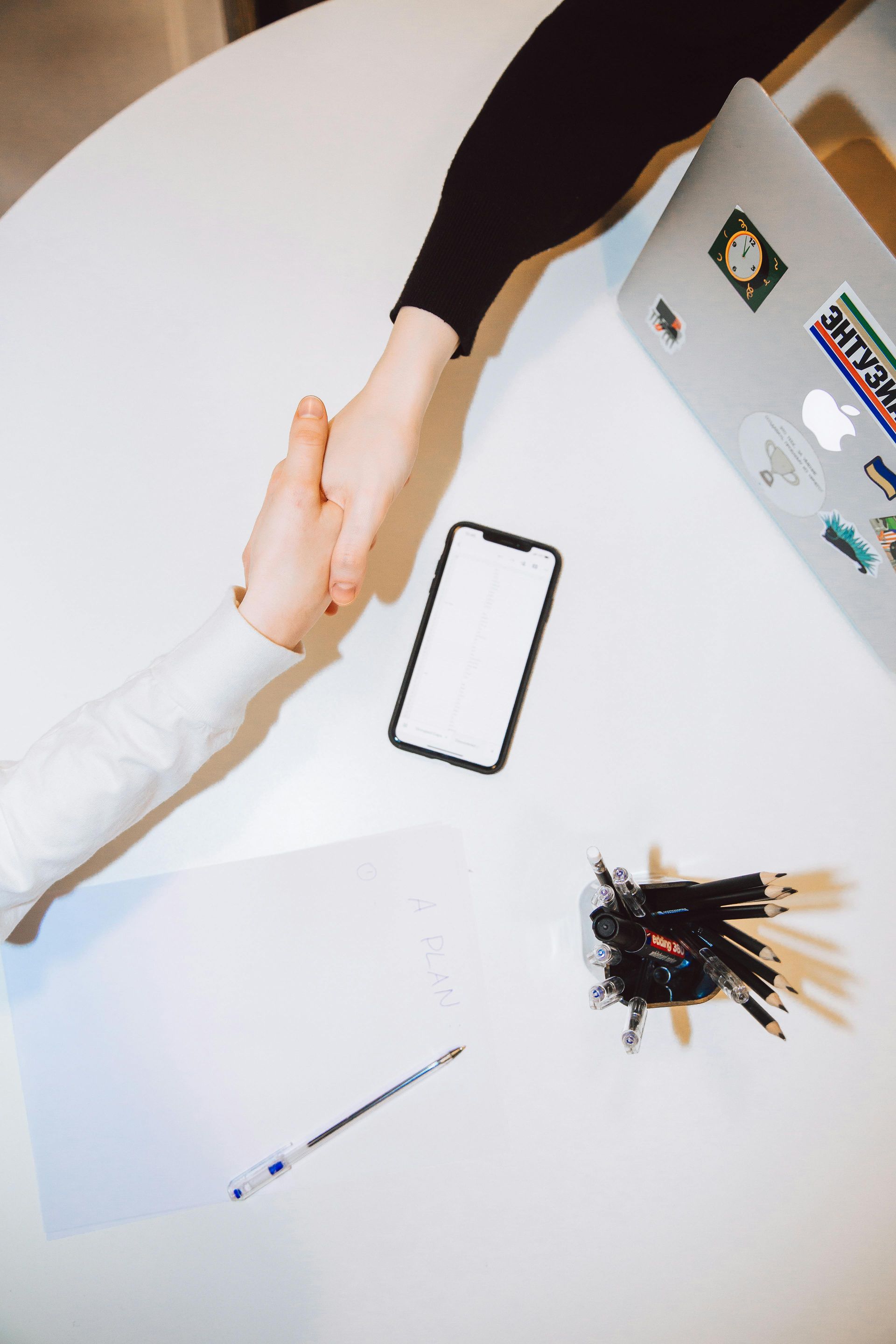 Two people are shaking hands over a table with a laptop and a cell phone.