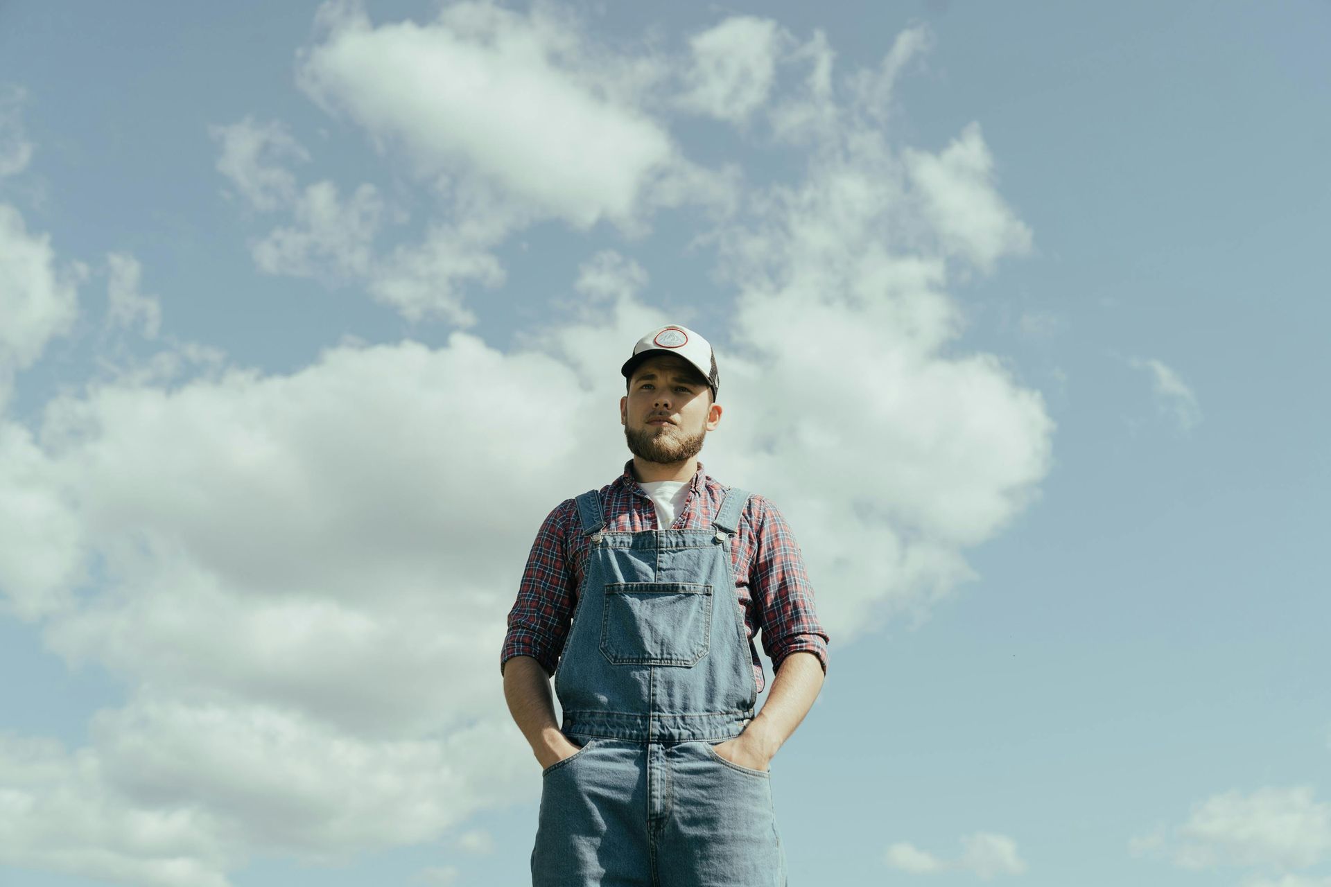 A man in overalls and a hat is standing in front of a cloudy sky.