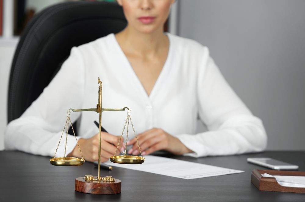 A Woman is Sitting at a Desk With Scales of Justice in Front of Her — Chris Trevor & Associates in Rockhampton, QLD