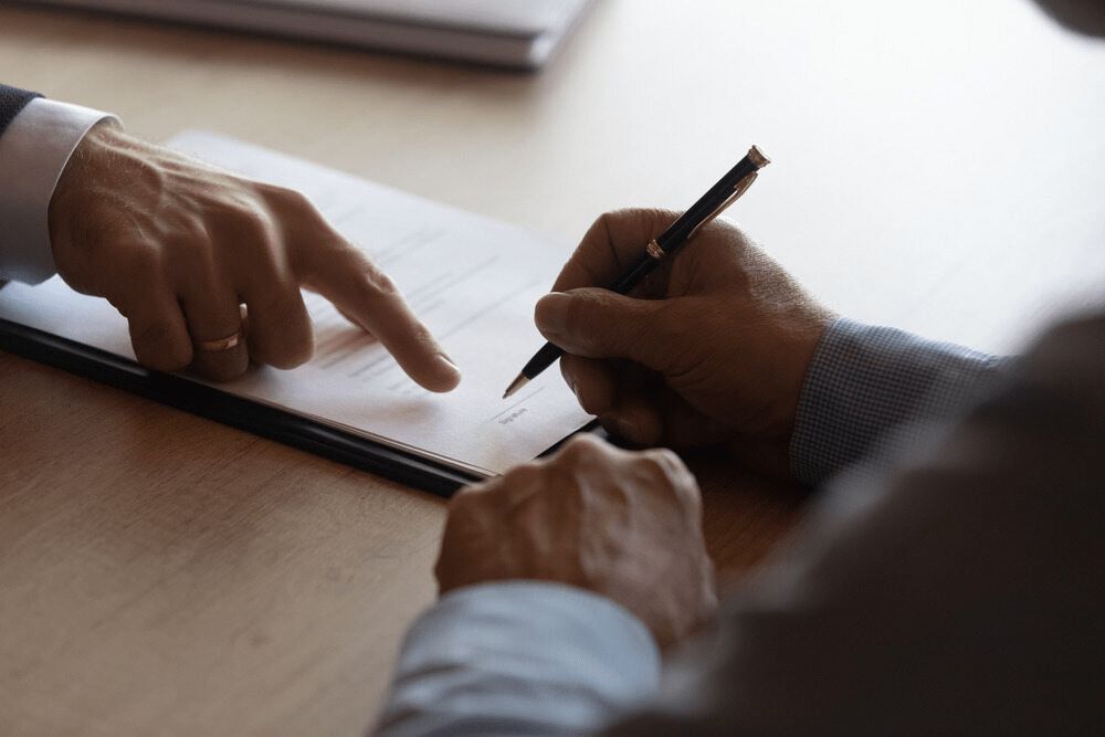 A Man is Signing a Document With a Pen While Another Man Points at It — Chris Trevor & Associates in Gladstone Central, QLD