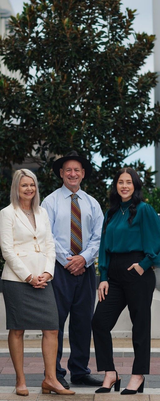 Three people standing outdoors: a woman in white, a man in a hat, and a woman in pink. They smile toward the camera.— Chris Trevor & Associates in Gladstone Central, QLD