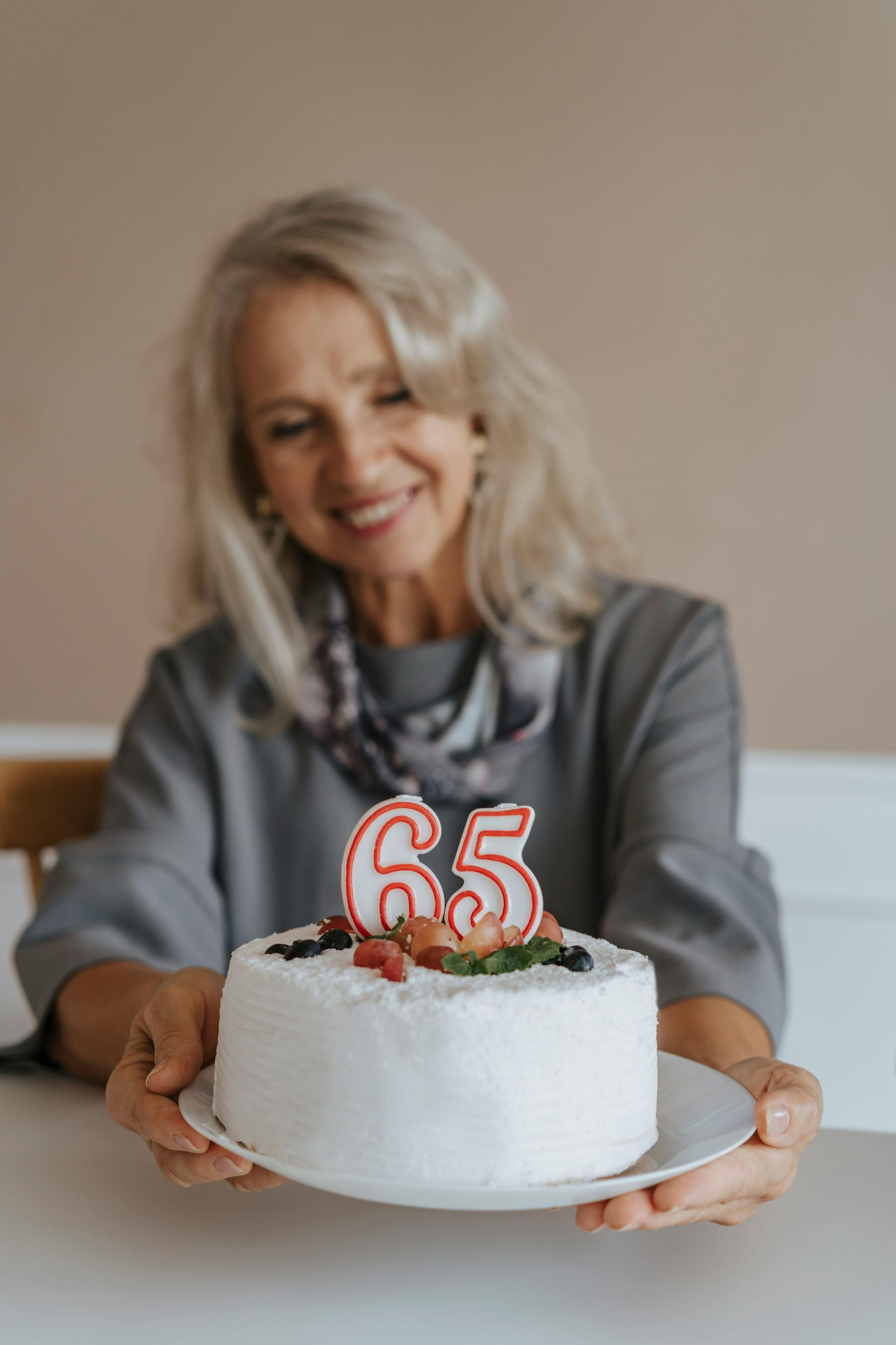 Woman holding a cake with a “65” candle. She smiles, sitting at a table.