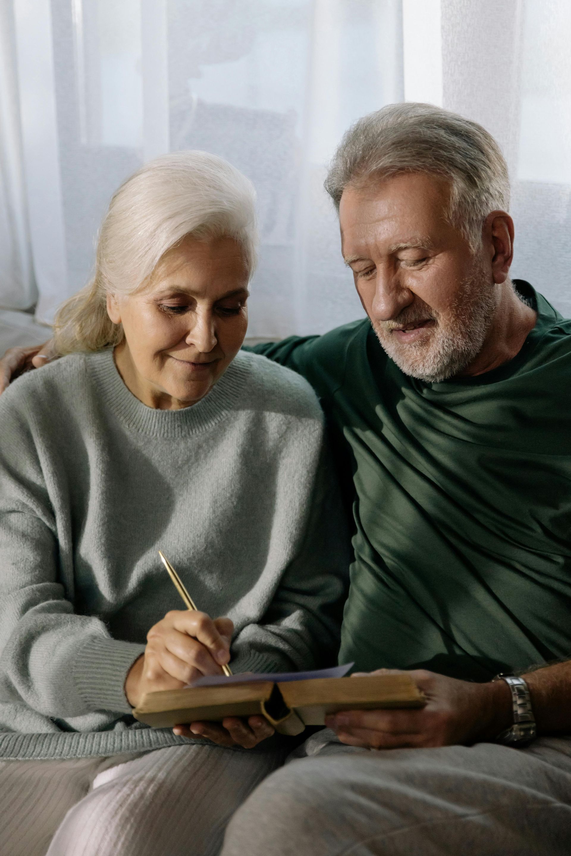 Elderly couple looking at a book; woman writes with a pen, man's arm around her.