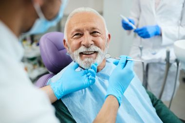 An elderly man is sitting in a dental chair while a dentist examines his teeth.