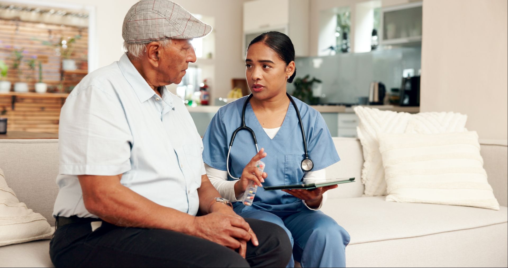 A home health aide in blue scrubs discusses information on a tablet with an older patient sitting on a couch.