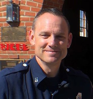 A man in a police uniform is smiling in front of a brick building.