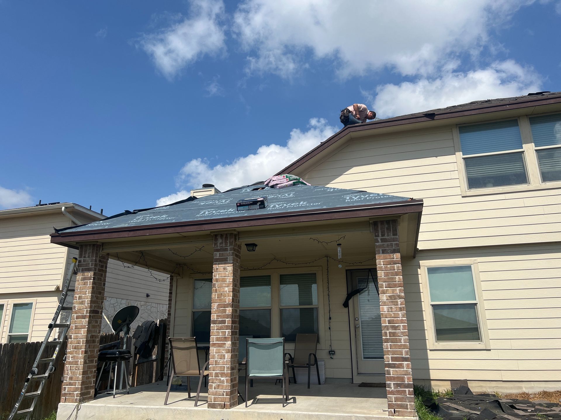 Roof repair on a house; a person on the roof under a blue sky, some shingles removed, a patio below.
