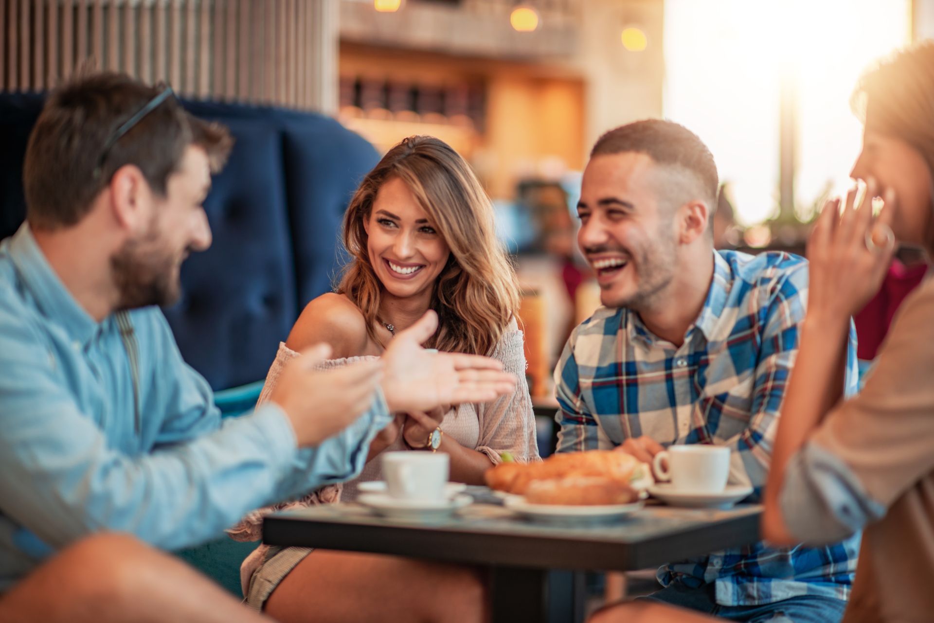Four people laughing and talking at a cafe table, enjoying food and drinks.