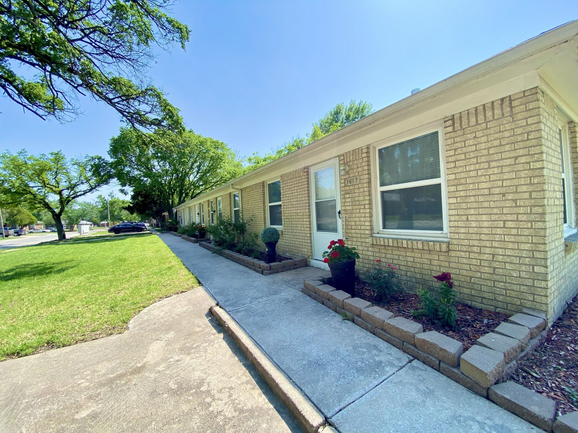 Row of tan brick apartment units with small flowerbeds along a sidewalk, under a bright blue sky.