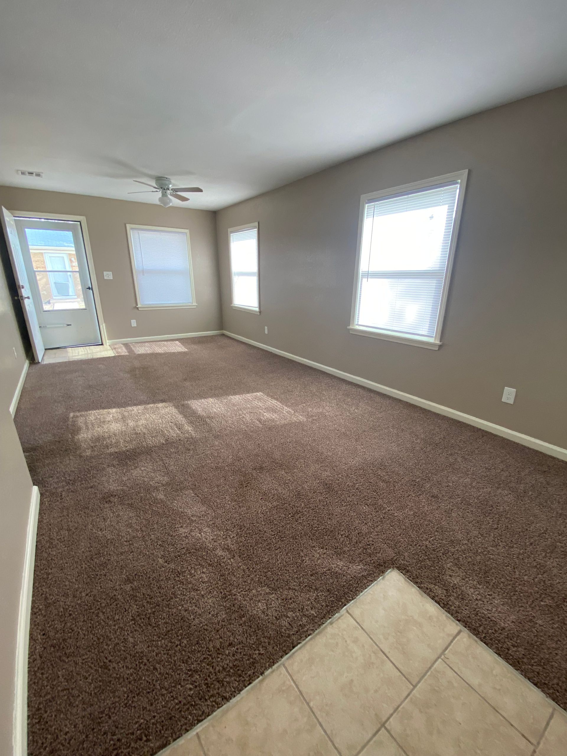 Empty living room with brown carpet, beige walls, and three windows.