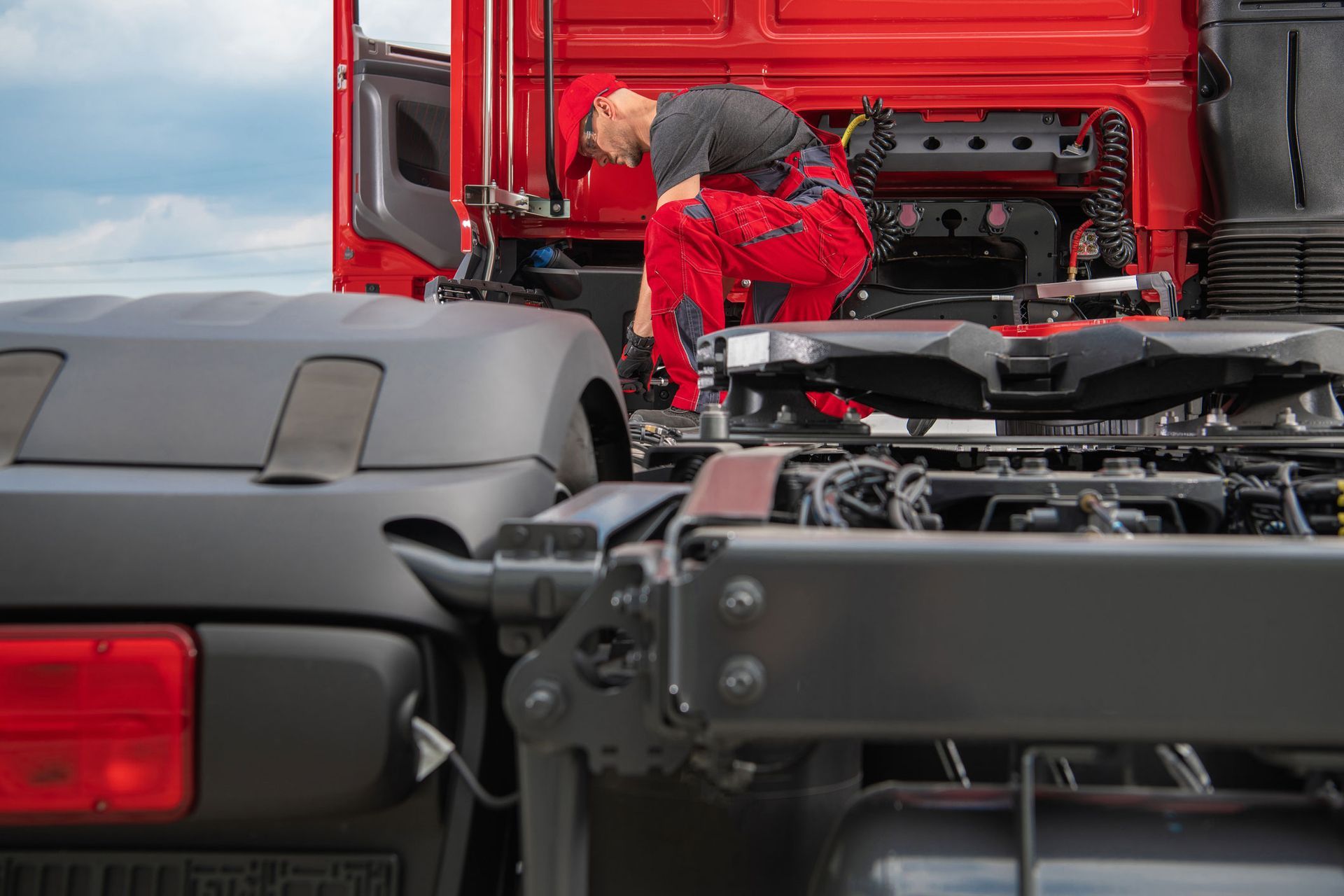 Person in red overalls inspecting the rear of a red semi-truck. Cloudy sky backdrop.