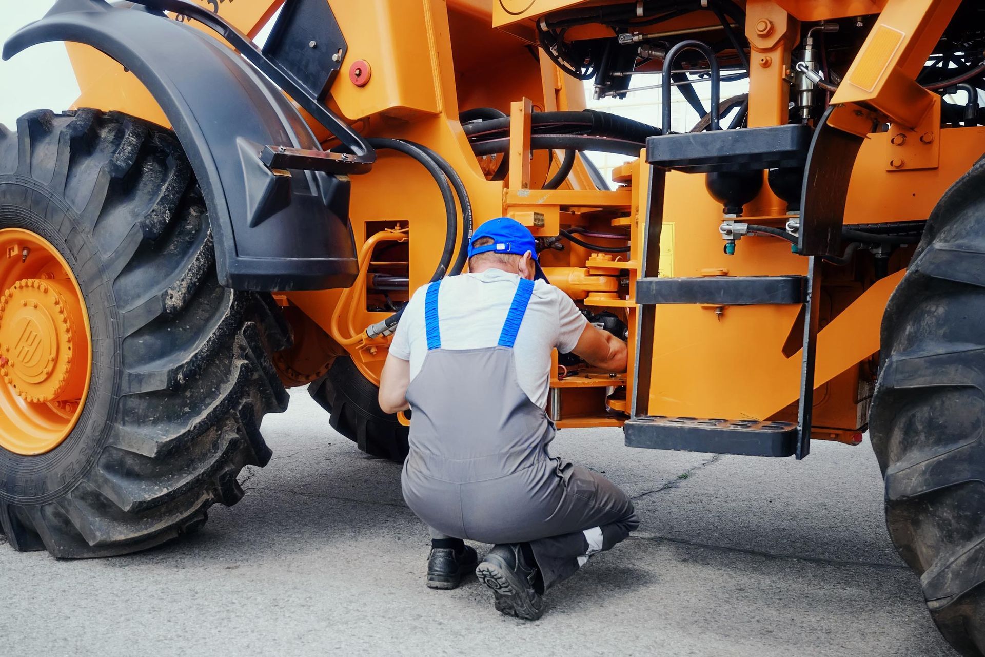 Mechanic in blue hat, overalls, inspecting an orange heavy-duty construction vehicle.