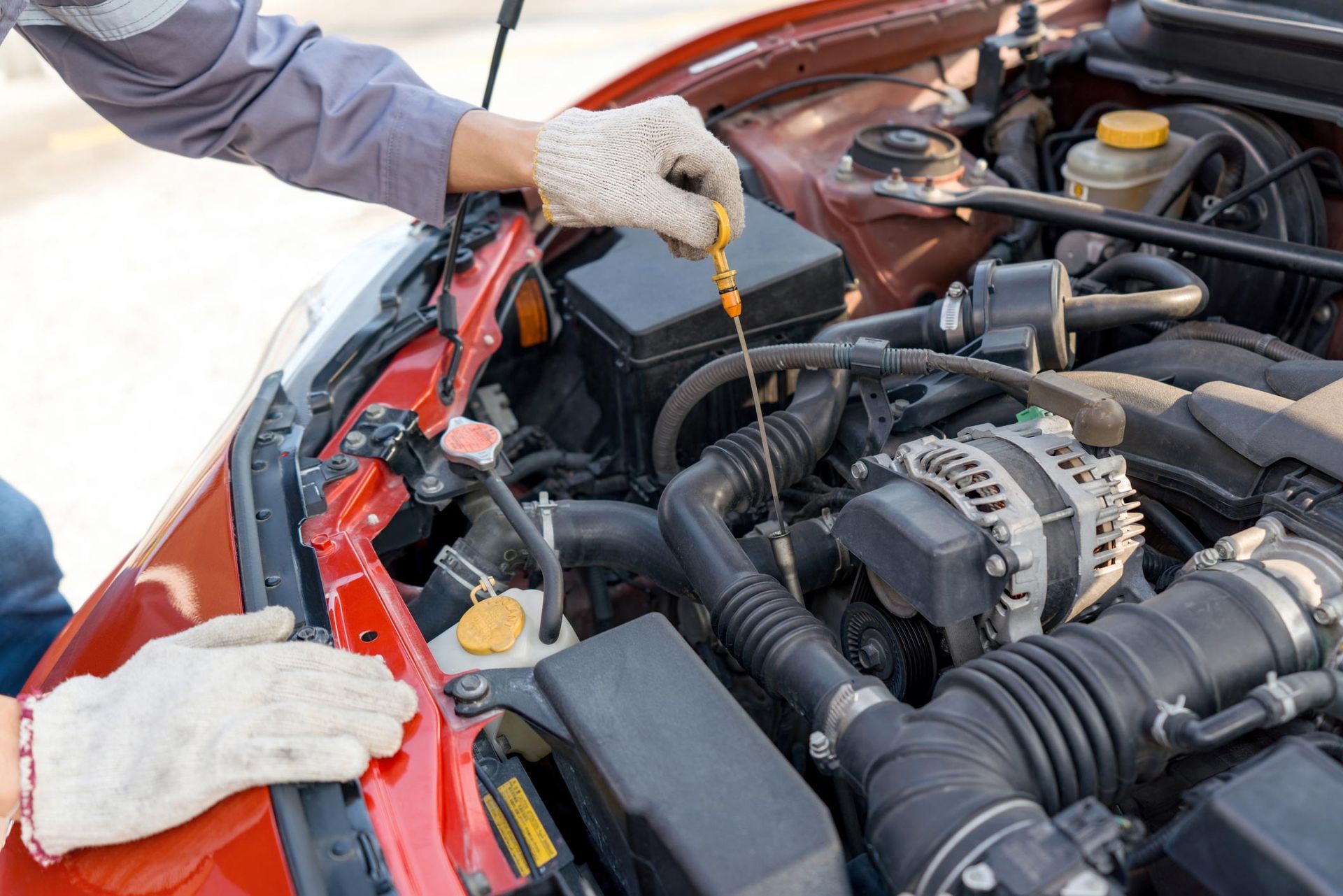 Person wearing gloves checking car's oil dipstick in a red engine bay.
