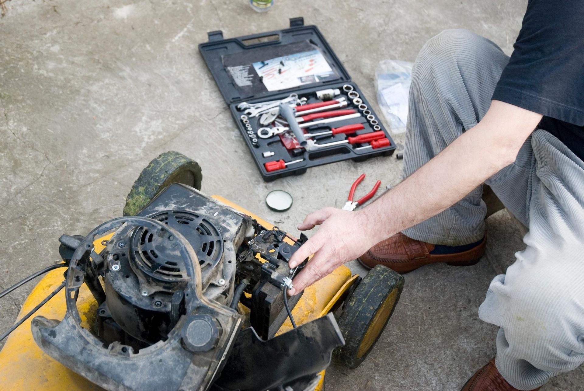 Person repairs a yellow lawnmower with tools from an open toolbox on concrete.