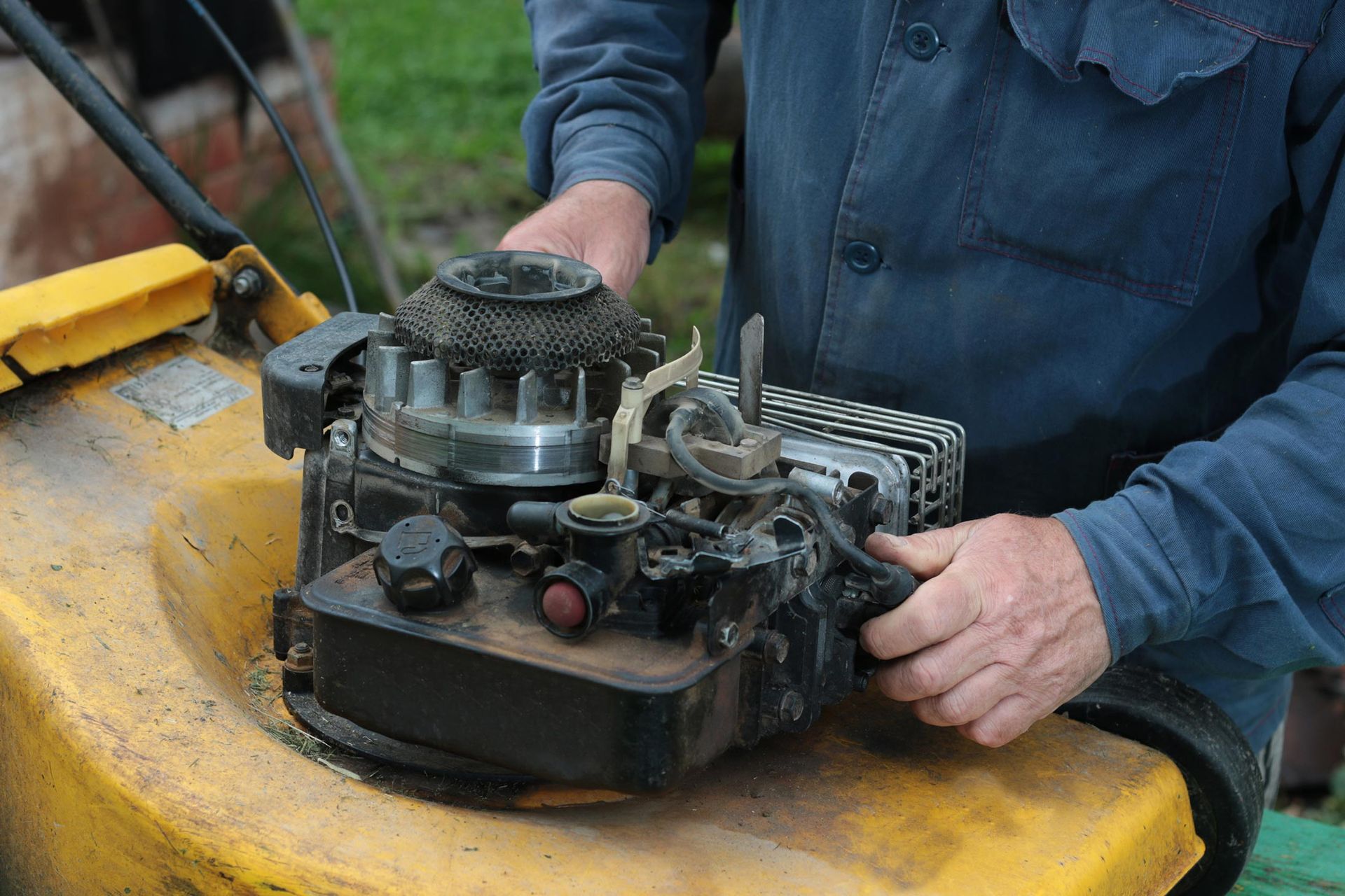 Person repairs a lawnmower engine; hands on parts. Yellow mower, blue shirt, outdoor setting.
