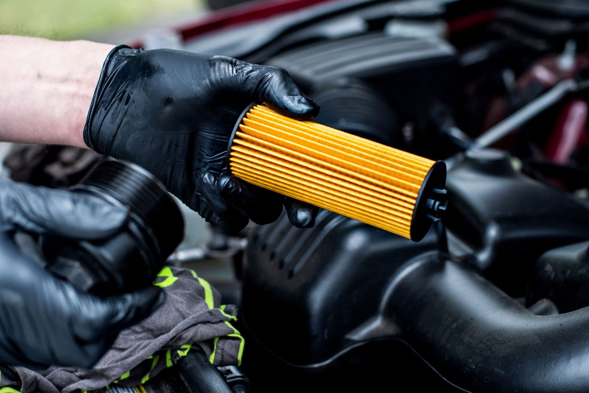 Hands wearing black gloves holding a yellow car oil filter near an engine.