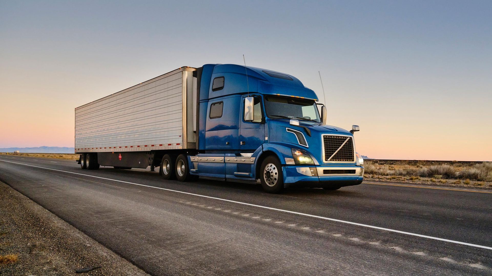 Blue semi-truck driving on a road with a white trailer, set against a sunset sky.