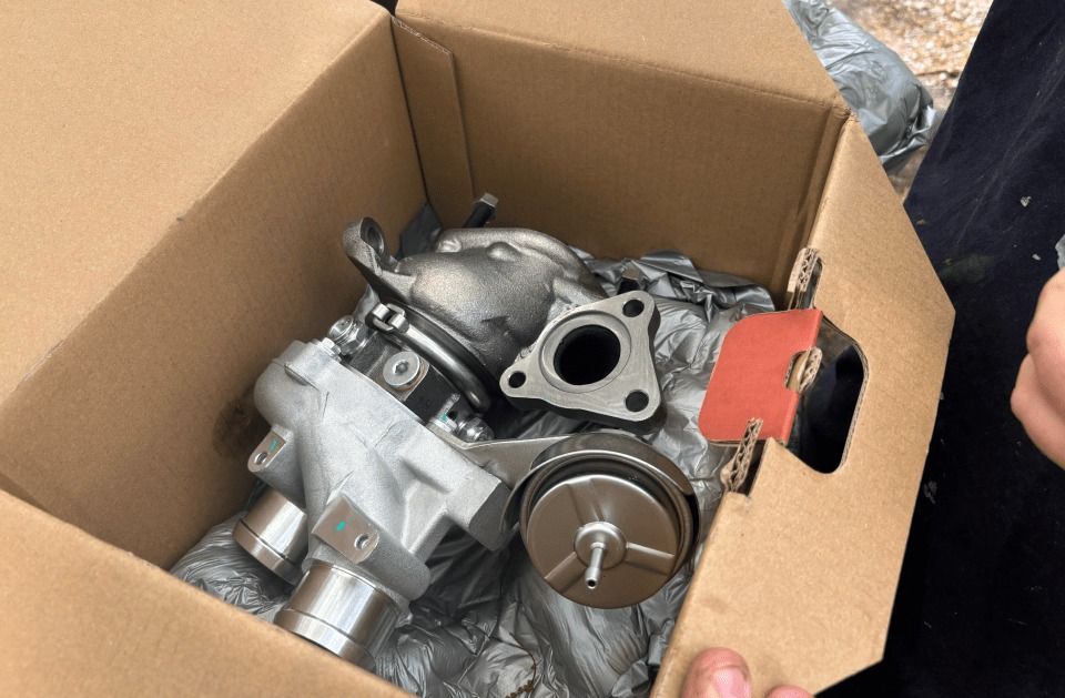 A new car turbocharger and related parts are in an open cardboard box, wrapped in plastic.