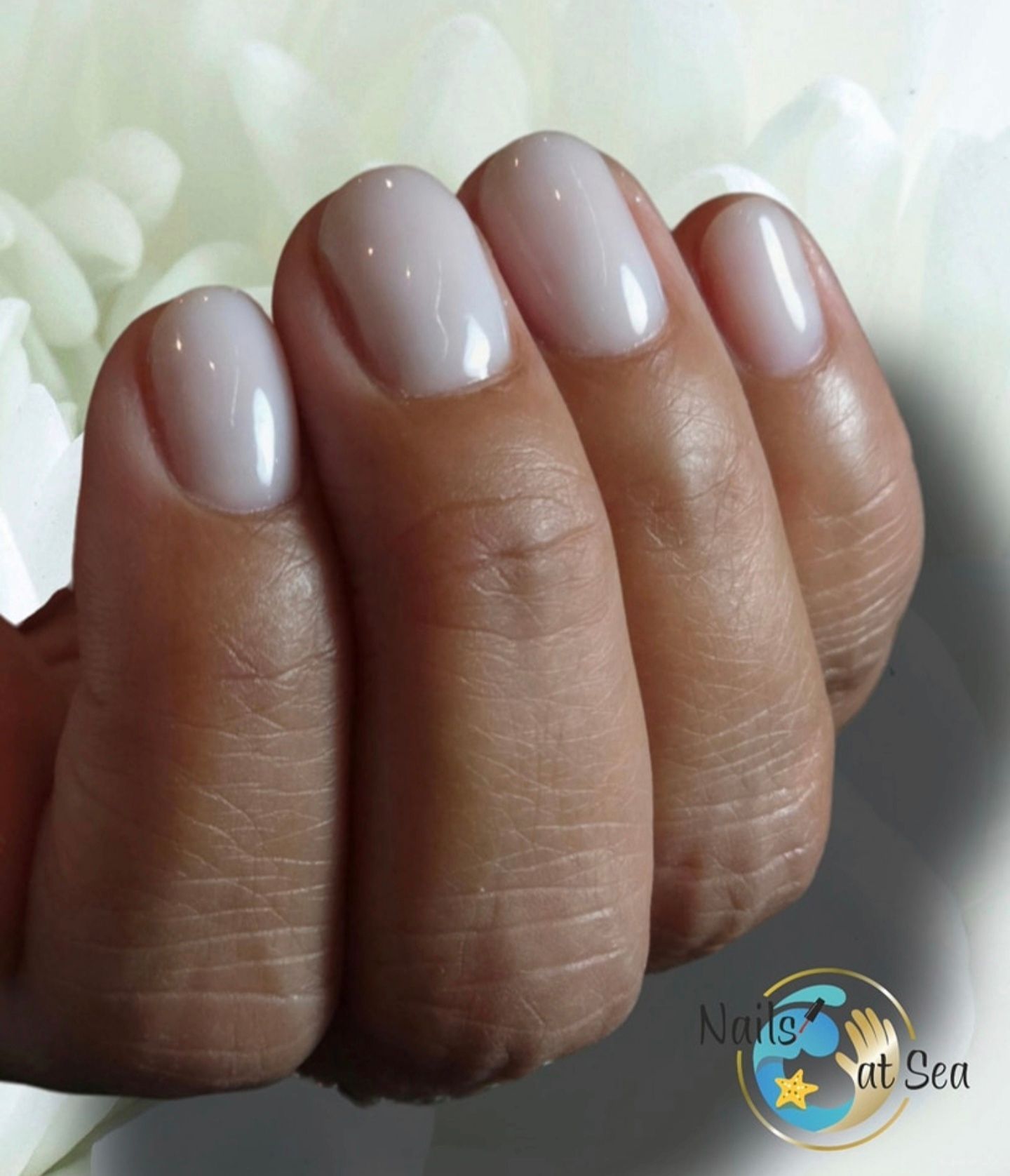 Een hand met korte, ronde, glanzende, gebroken witte nagels tegen een witte bloemenachtergrond.