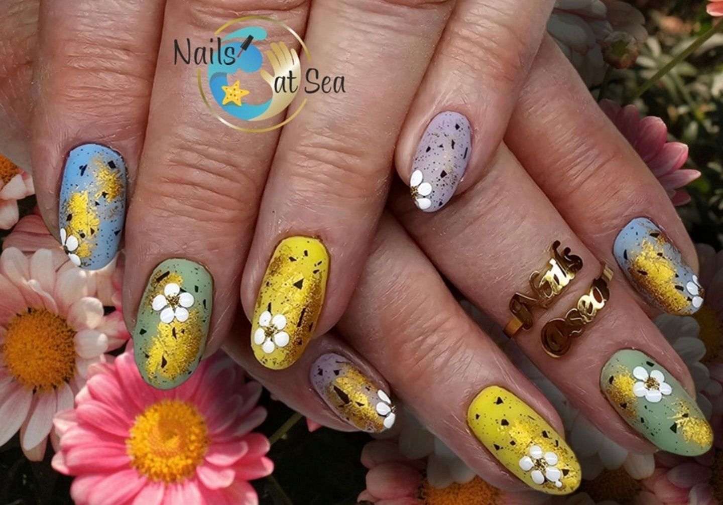 Handen met amandelvormige nagels, versierd met goudfolie, witte bloemaccenten en gele, blauwe en saliegroene nagellak.