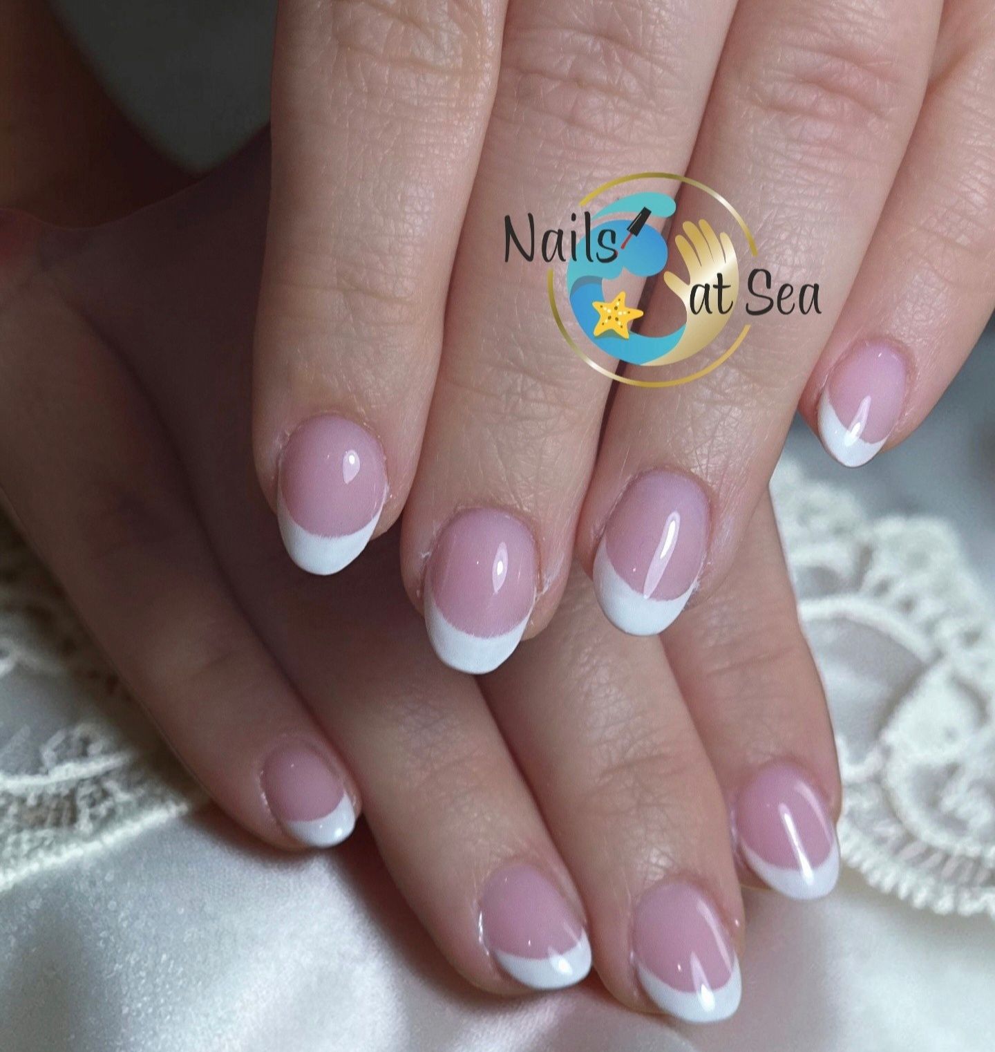 Handen met een klassieke Franse manicure, een nude basis en helderwitte nagelpunten, afgebeeld tegen een delicate kanten achtergrond.