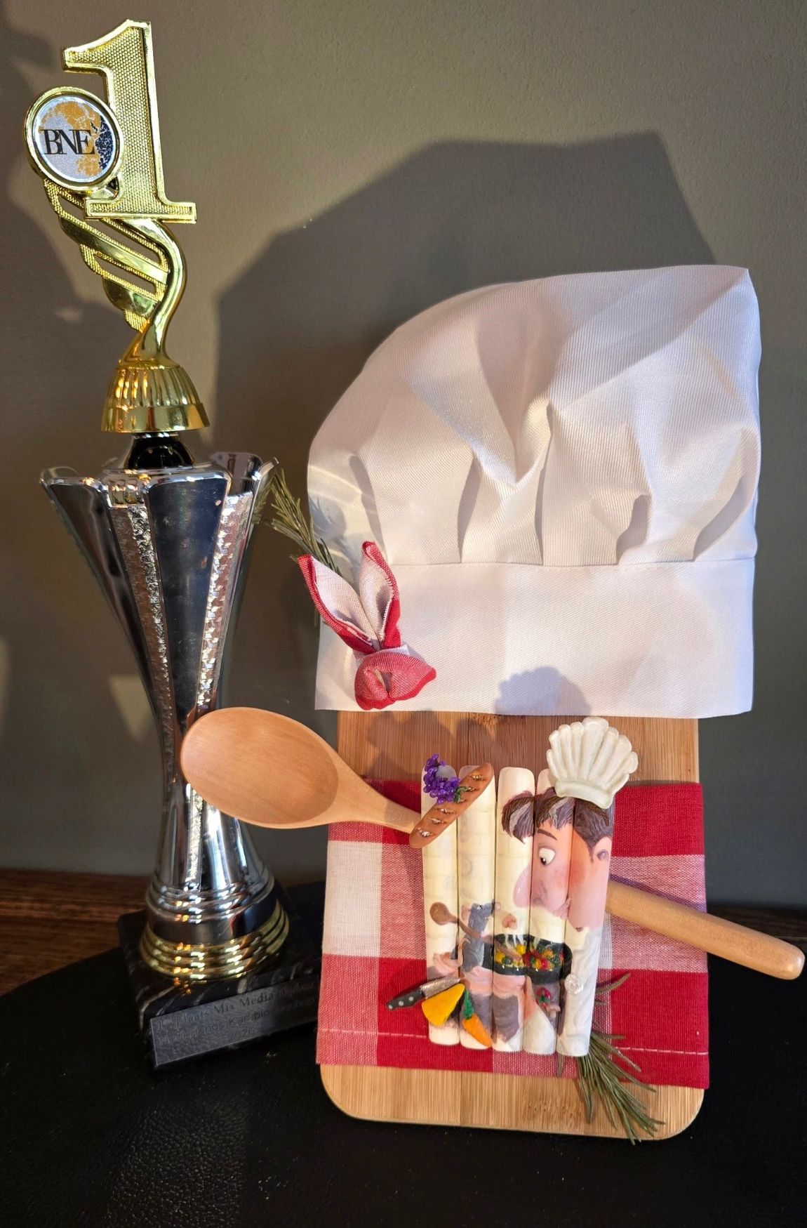 Naast een houten snijplank, versierd met een koksmuts, rood geruite stof en keukengerei, staat een trofee voor de eerste plaats.