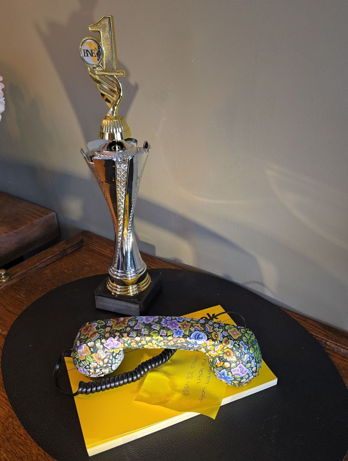 Een trofee staat achter een telefoonhoorn met bloemenmotief, die rust op een geel notitieblok op een zwarte ronde tafel.