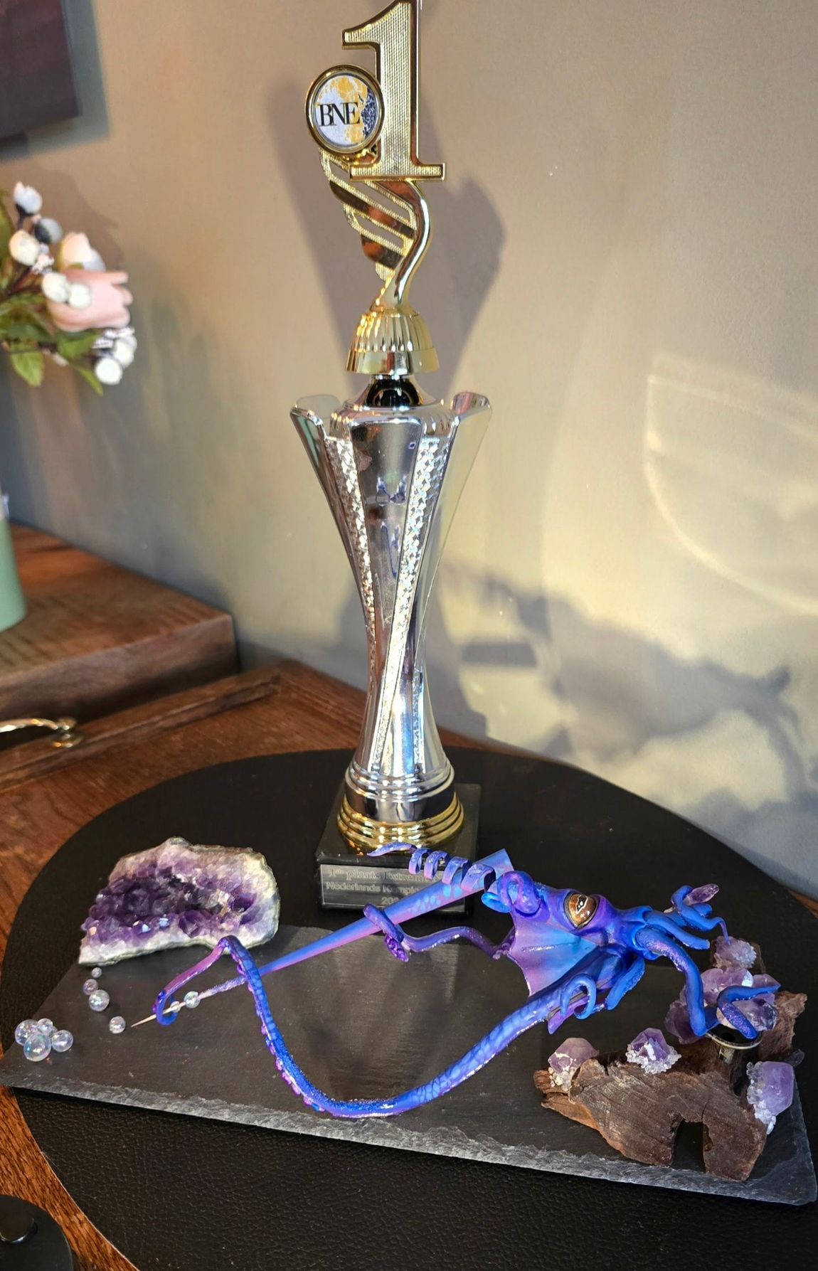 Een met goud bekroonde trofee, een ruwe amethistgeode en een blauw draakje, gerangschikt op een zwarte leistenen ondergrond.