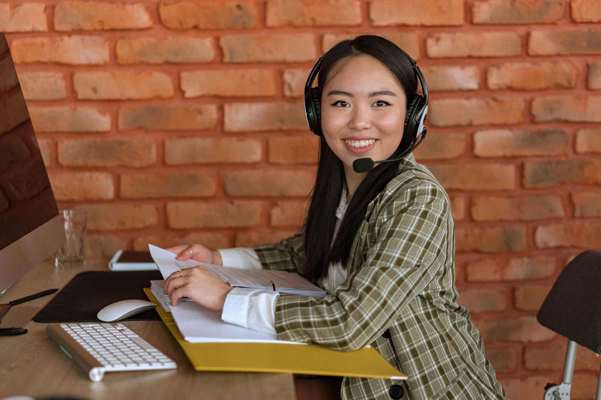 Person wearing headphones and a plaid shirt working at a desk with papers and a laptop, smiling in a brick-walled room