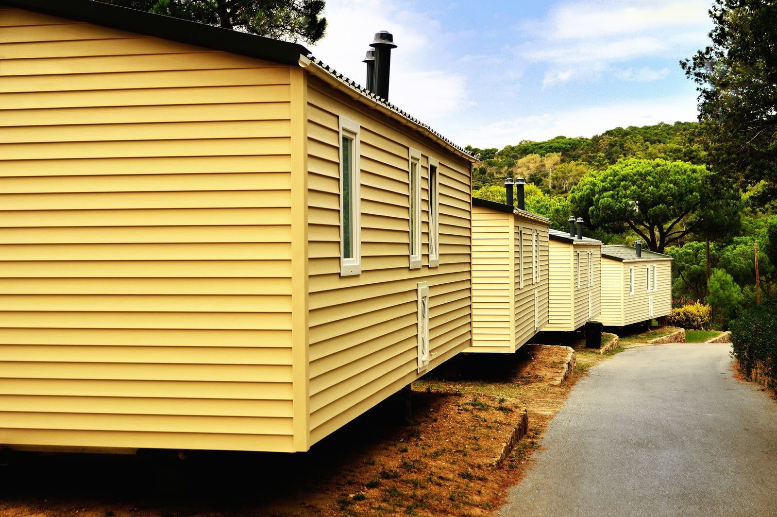 Row of cream-colored cabins along a paved path beside green trees under a blue sky