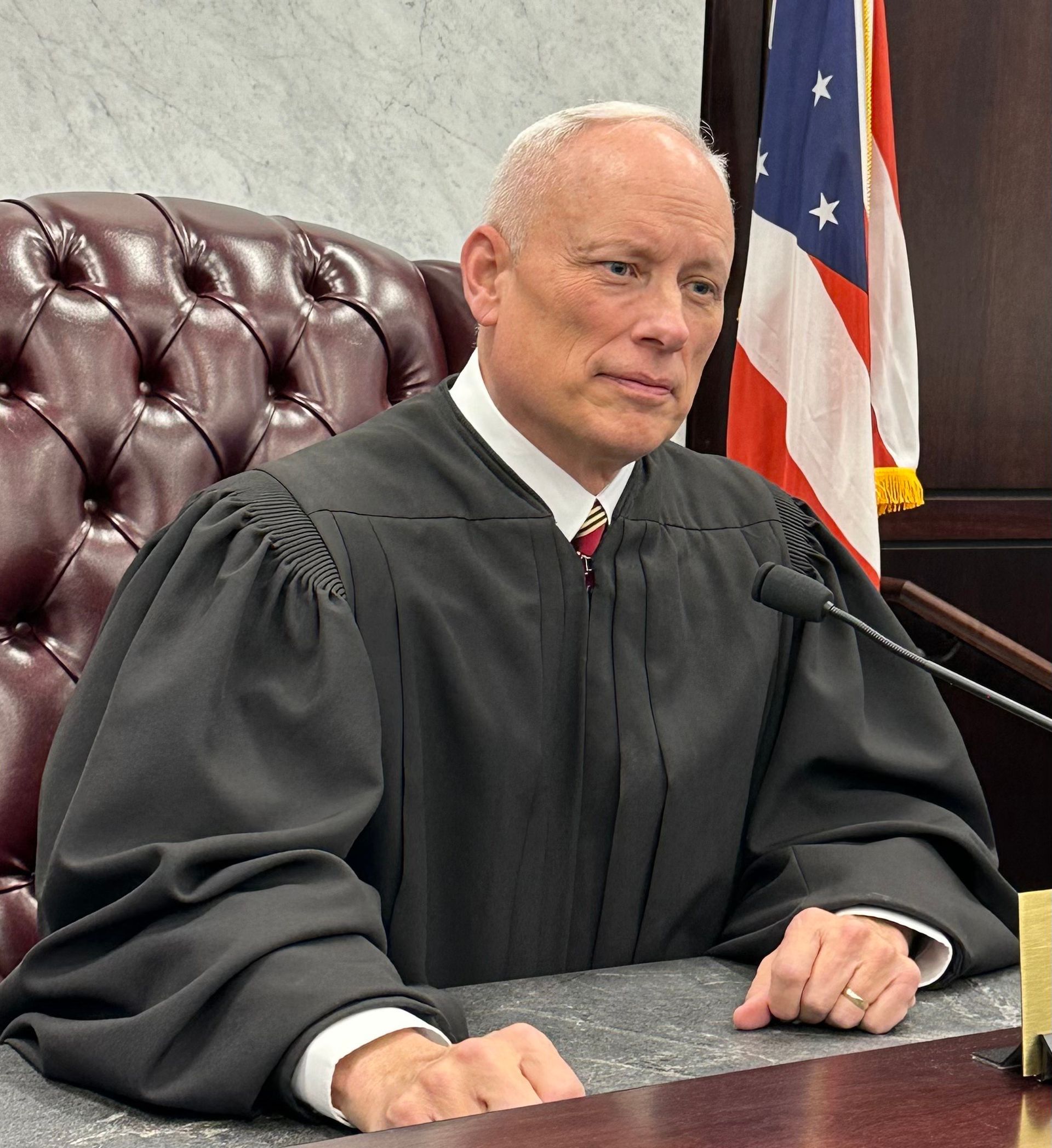 Judge Gary A. Loxley seated at a desk, wearing a robe, in a courtroom with an American flag behind him.
