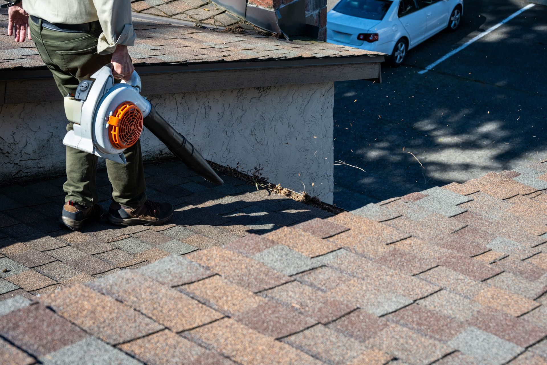 Person using a leaf blower to clear debris from a shingled roof next to a parking lot.