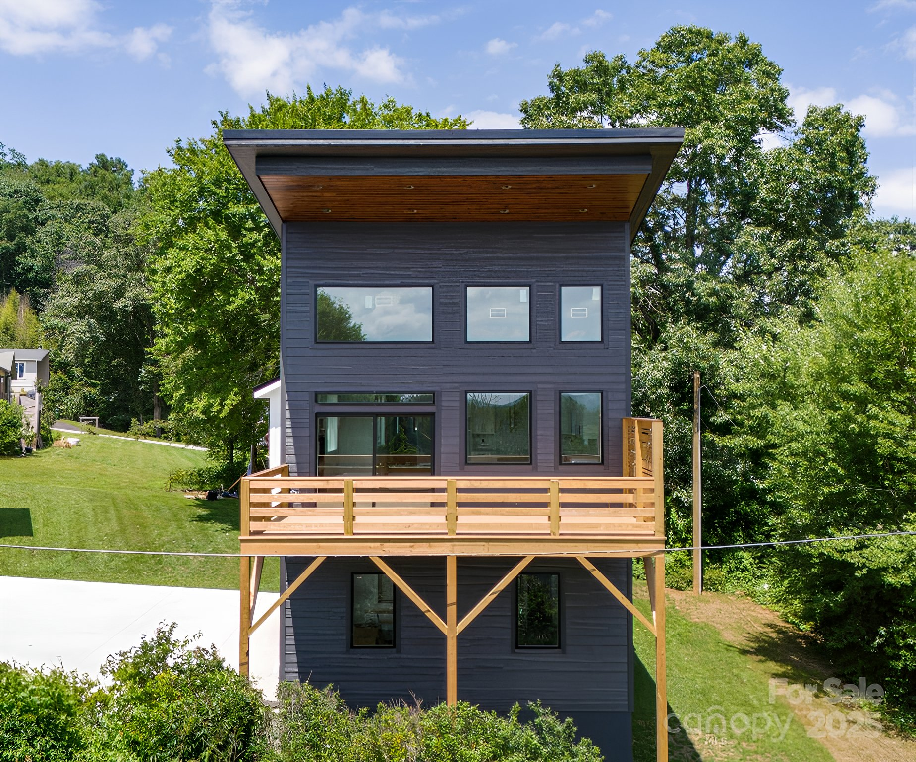 Modern, two-story house with dark gray siding, wooden deck, large windows, and a flat roof, surrounded by trees.