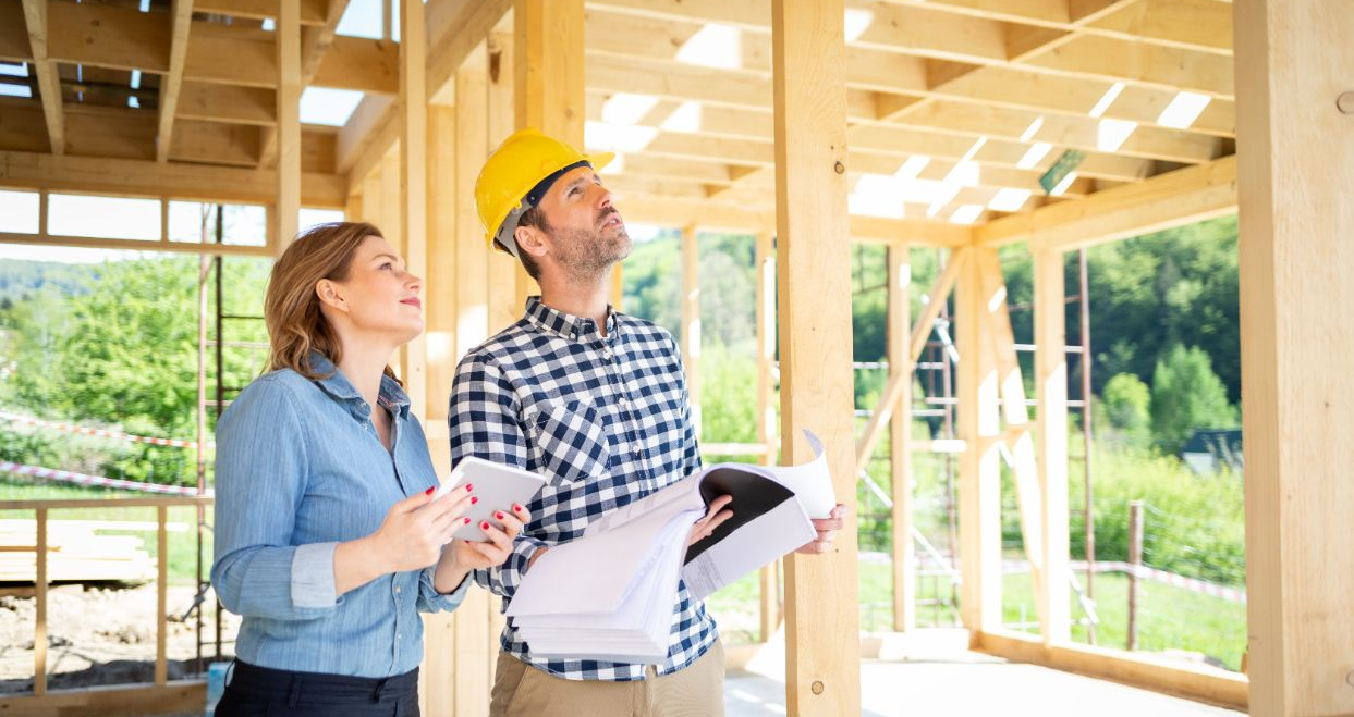 A man in a hard hat and woman examine blueprints inside a wooden house frame, surveying construction.