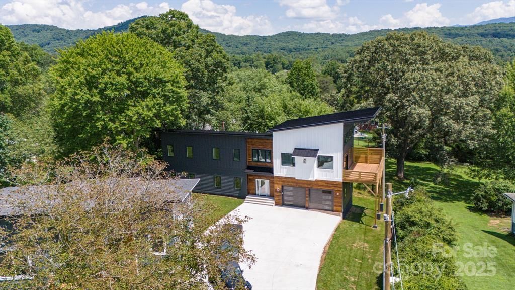 Modern house with black and white exterior, two-car garage, and a long driveway surrounded by lush trees and a mountain background.