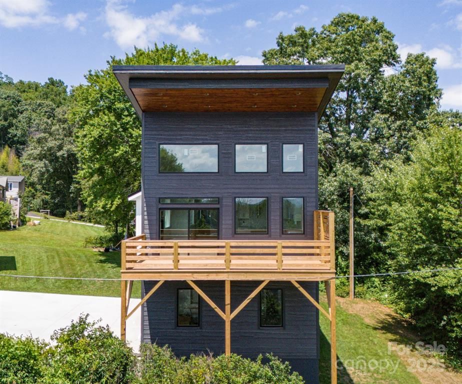 Modern, three-story house with a wooden deck and dark gray siding surrounded by trees and green grass.