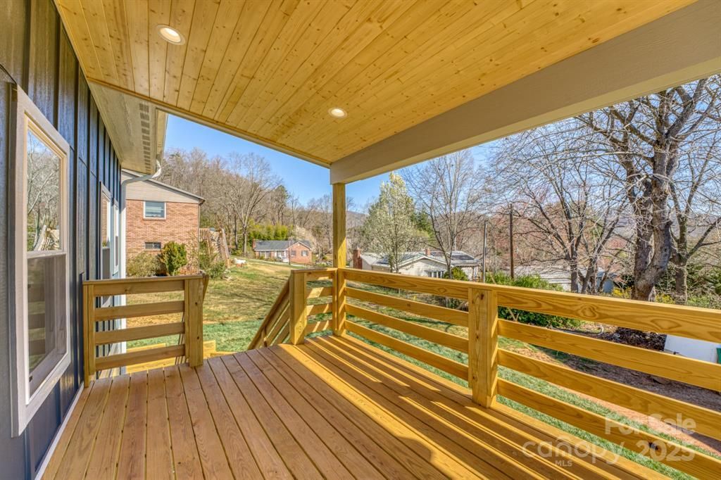 Wooden deck with railing, under a covered porch with blue siding. Overlooks yard with trees.