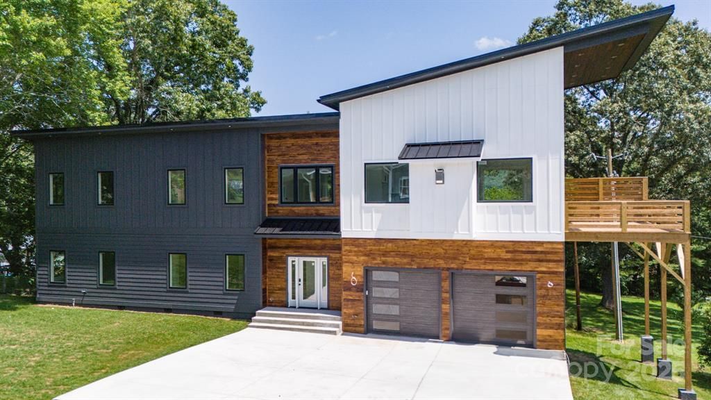 Modern two-story house with dark gray siding, wood accents, and a sloped roof. Garage with glass doors and a wooden deck.