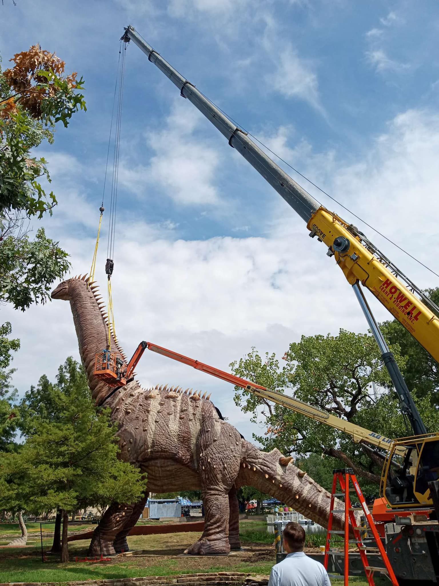 A large dinosaur sculpture being lifted by a crane. The dinosaur is brown and textured, in an outdoor setting with a blue sky.