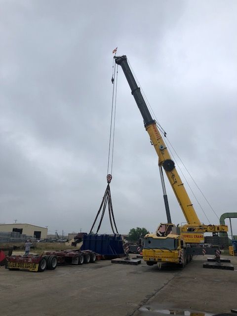 A yellow crane lifting a large, dark-colored object, likely for transport onto a flatbed trailer, under a cloudy sky.