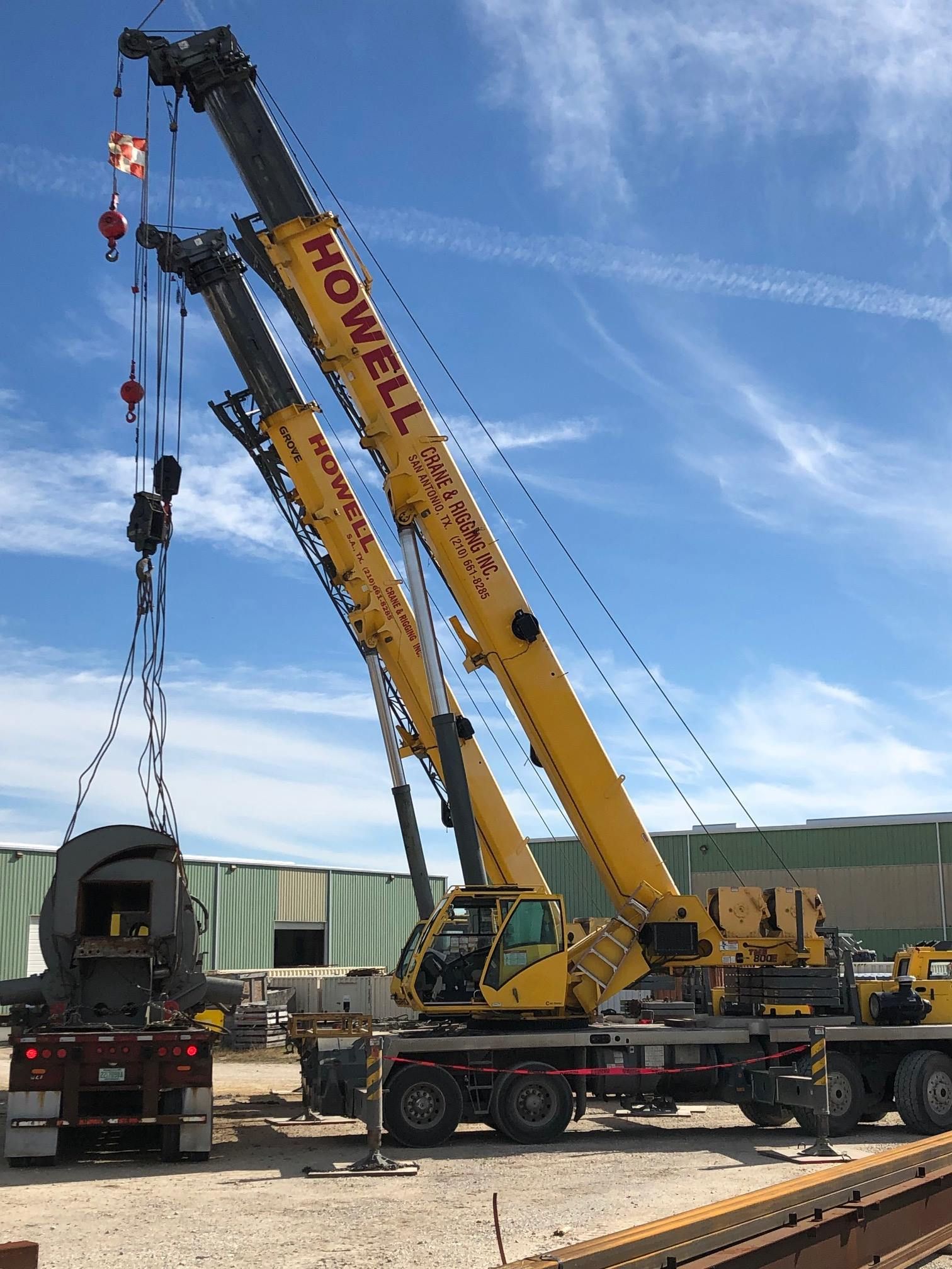 Two yellow Howell cranes lifting a large, dark object from a flatbed truck under a blue sky.