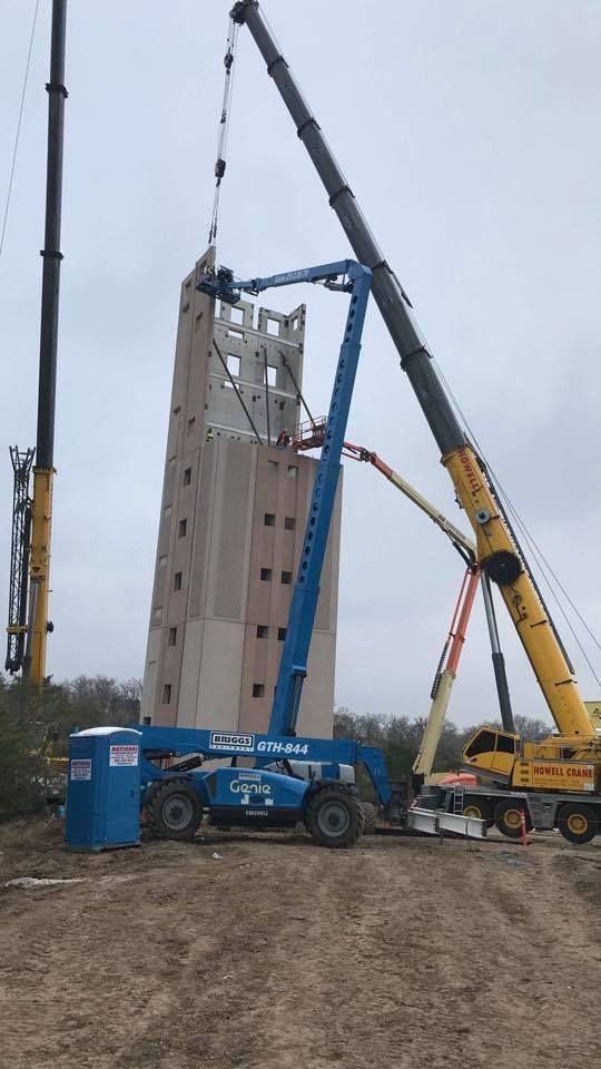 Two cranes lift a tall, precast concrete structure, with a blue lift truck positioned nearby. Construction site on an overcast day.